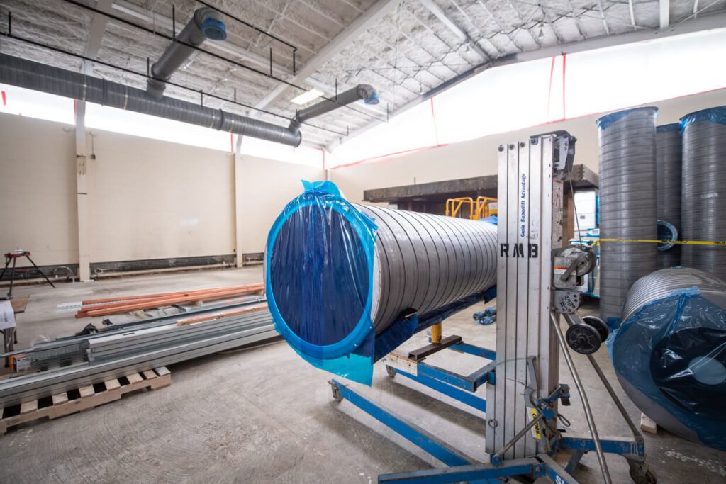 Large metal ducting wrapped in blue plastic sits on a lift in a spacious, unfinished building with exposed ceiling and materials like pipes and pallets scattered on the concrete floor.