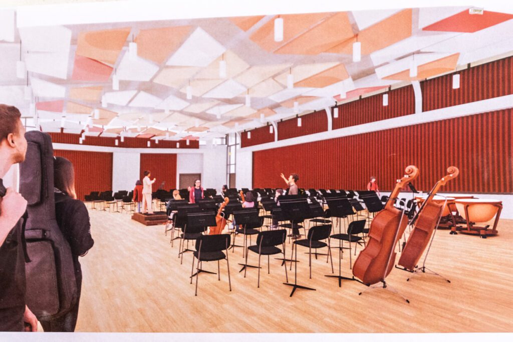 A modern orchestral rehearsal room with musicians holding their instruments, empty music stands, cellos and timpani in the corner, and geometric ceiling panels above light wood floors and red wall accents.