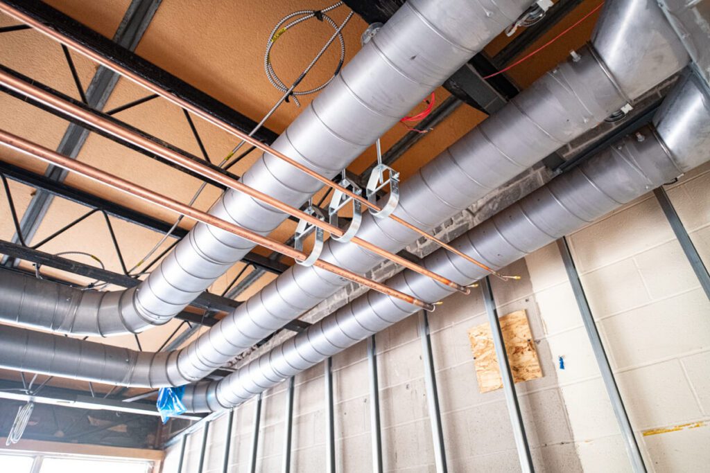 Exposed metal air ducts and copper pipes run along a ceiling with visible beams and wiring in a building under construction. Unfinished walls and insulation are also visible.