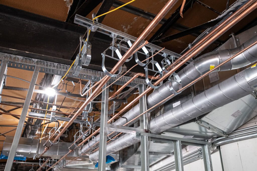 Exposed pipes and ducts run along an unfinished ceiling in a building under construction, with metal framing, insulation, and visible wiring.