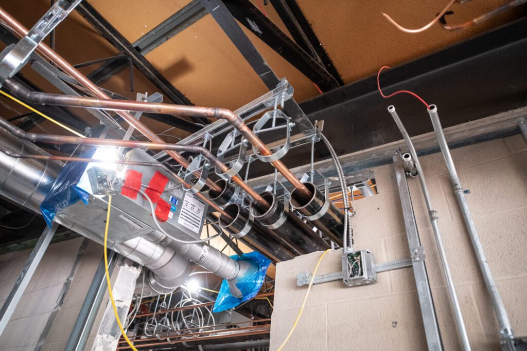 View of exposed pipes, conduits, and ductwork on a ceiling under construction, with some insulation, wiring, and metal framing visible against a partially finished wall.