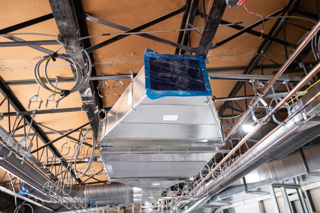 Large metal HVAC ductwork is installed on a ceiling with exposed beams, wires, and pipes in an unfinished building interior. Blue plastic covers one duct opening.