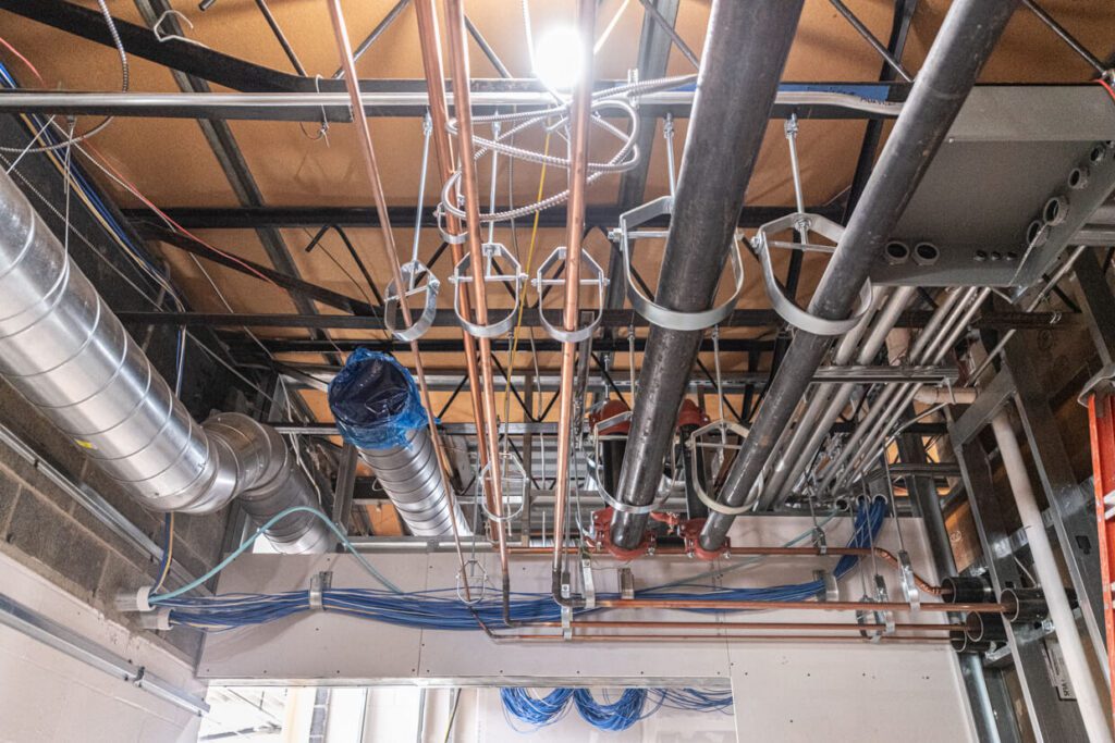 Exposed ductwork, pipes, and cables run along the unfinished ceiling of a building under construction, with metal framing and insulation visible.