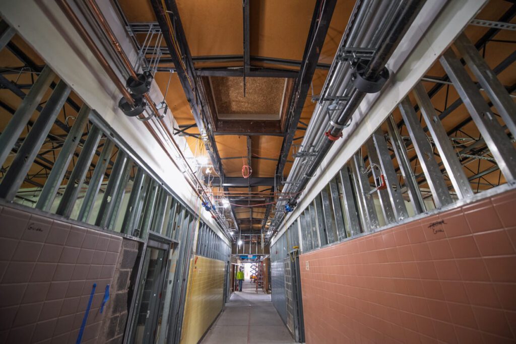 A hallway under construction with exposed metal framing, visible pipes and wiring on the ceiling, and partially tiled walls in yellow and brown. Bright lights illuminate the work area in the distance.