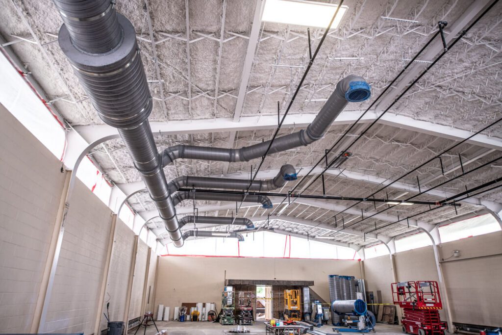 Large industrial room with exposed ductwork on the ceiling, construction equipment, and materials scattered on the floor, including ladders, pallets, and rolls of insulation, under bright overhead lighting.