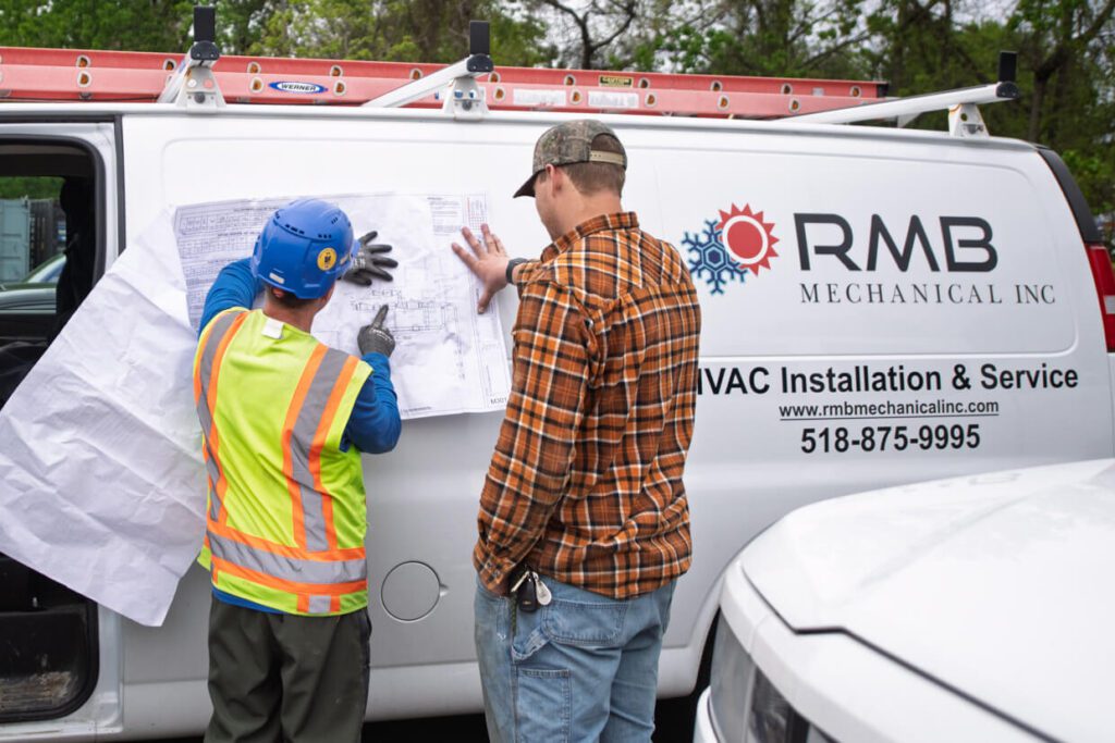 Two workers review blueprints taped to a white van labeled RMB Mechanical Inc. One worker wears a hard hat and vest, the other a plaid shirt and cap. A ladder is mounted on the van roof.