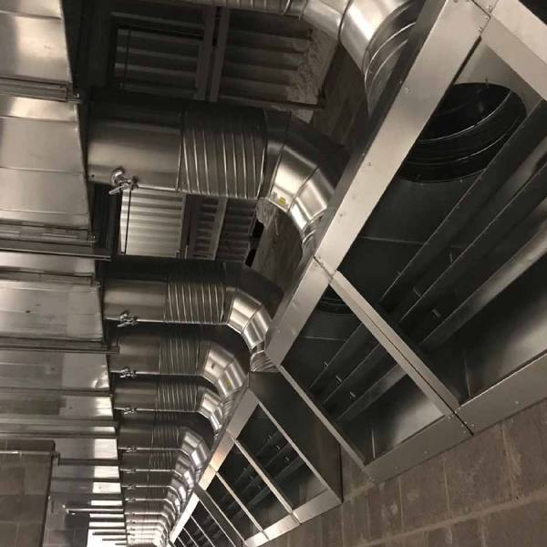 Industrial metal air ducts and vents mounted on a ceiling, part of the building’s HVAC system, with several rectangular openings along the side, inside a building with concrete block walls.
