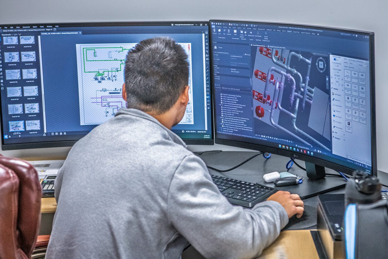 A person sits at a desk using two large monitors, one displaying technical diagrams related to Building Information Modeling and the other showing a 3D model of a mechanical system. Various office items are on the desk, including a keyboard and water bottle.