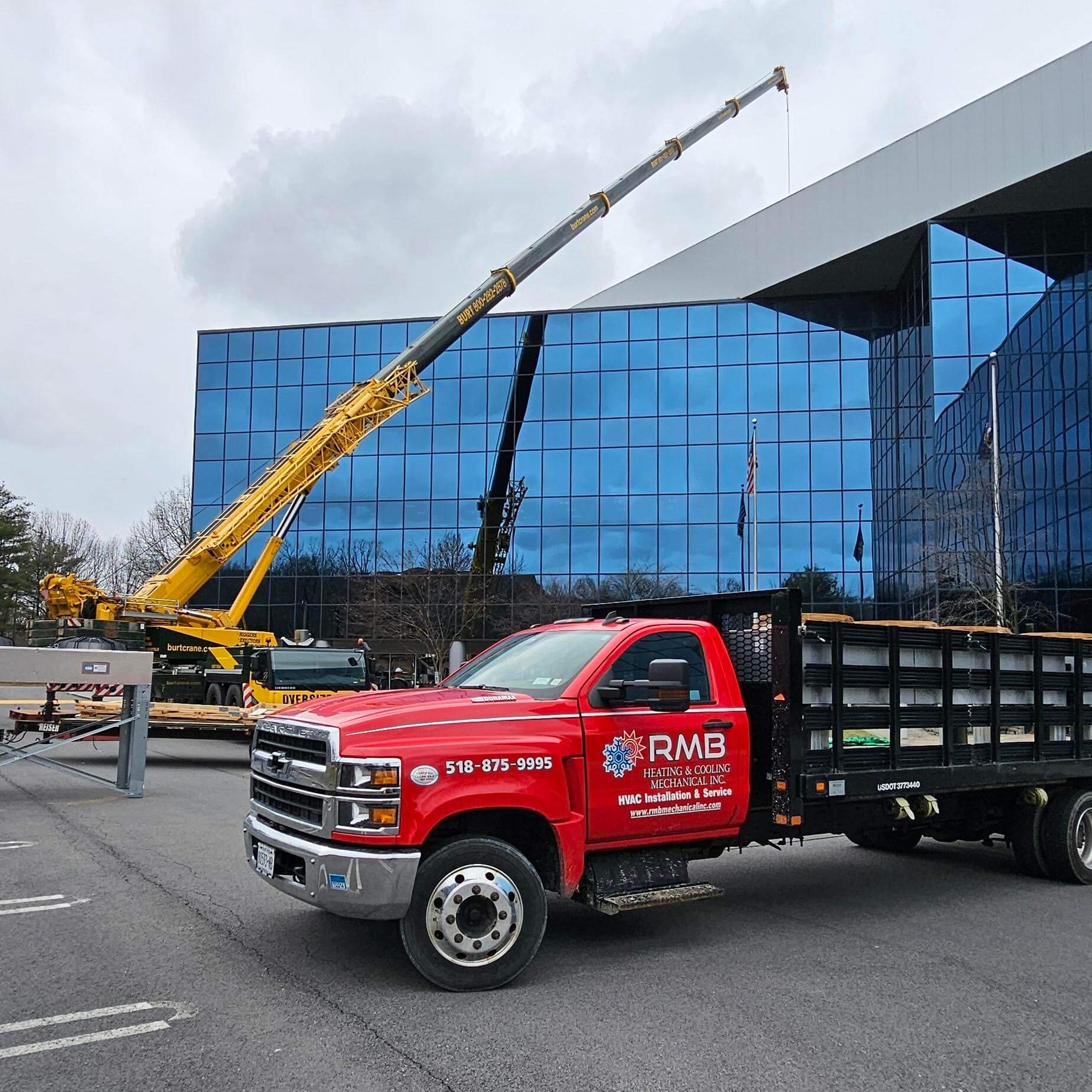 A red RMB truck is parked in front of a modern glass building, while a large yellow crane lifts materials nearby on a cloudy day. For more details about our services or to get in touch, please contact us.