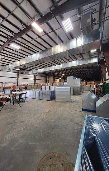 Large industrial warehouse interior with metal ducts, work tables, tools, and equipment scattered throughout. Overhead lights illuminate the spacious area with high ceilings and exposed steel beams.