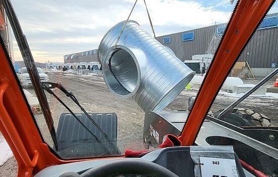 A large metal ventilation duct is being lifted by machinery outside a building, viewed from inside a vehicle cab on a construction site.