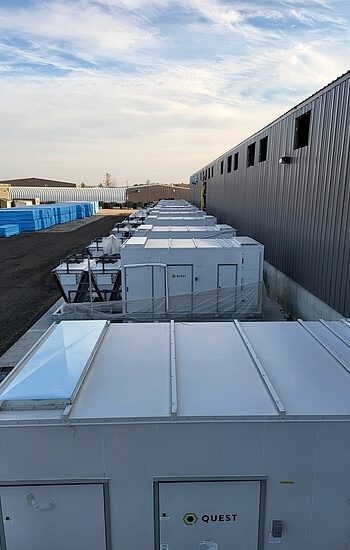 Rows of white, modular container units are lined up beside a long gray industrial building under a blue sky with light clouds. Blue containers are stacked in the distance.