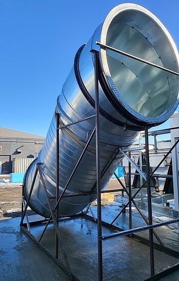 A large, shiny metal industrial duct is supported by a black metal frame outdoors on a concrete surface, with buildings and blue sky in the background.