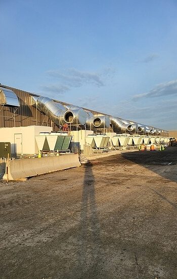 Large industrial building with multiple metal ventilation ducts and HVAC units along the exterior wall, set under a clear sky. Shadows and construction cones are visible on the ground.