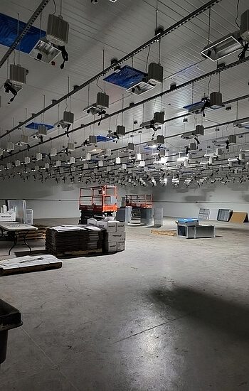 A large indoor room with many overhead grow lights, stacks of materials, carts, and construction equipment, suggesting a facility being prepared for indoor agriculture or horticulture.