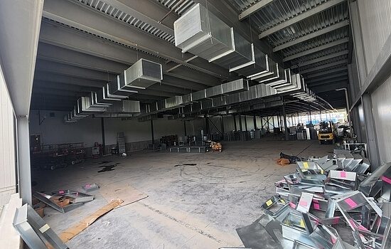 Large, unfinished warehouse interior with exposed metal beams, HVAC ductwork on the ceiling, scattered construction materials, and a forklift in the background. The space appears to be under construction or renovation.