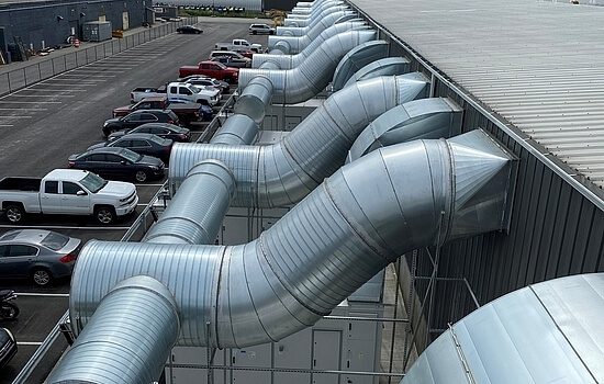 Large metal ventilation ducts emerge from a buildings exterior wall, extending over a row of parked cars in a parking lot beside an industrial facility.
