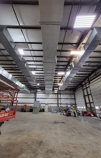 Large industrial warehouse interior with exposed ceiling ductwork, bright overhead lights, a few workers in safety vests, and equipment scattered around the mostly empty concrete floor.