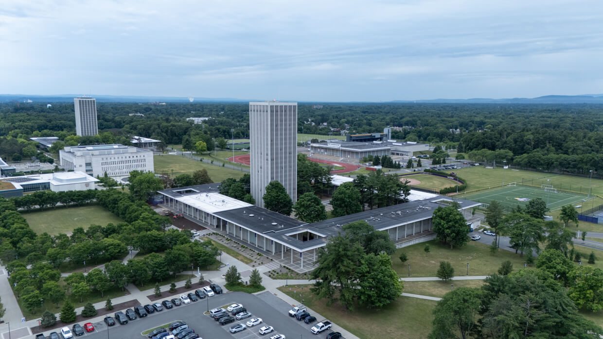 Aerial view of a university campus with tall buildings designed using Building Information Modeling, athletic fields, parking lot, and surrounding greenery under a cloudy sky.