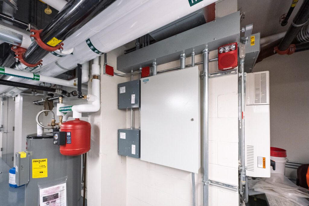 A utility room with electrical panels, wires, a red pressure tank, pipes, control boxes, and a water heater against a white wall. The area is organized and industrial in appearance, reflecting modern Building Information Modeling standards for efficient facility management.