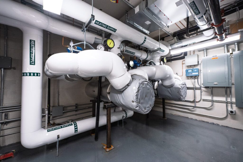 Large insulated pipes labeled Heating Supply with gauges, valves, and control panels are installed in a clean mechanical room with gray floors and exposed ceiling pipes, showcasing the precision planning enabled by Building Information Modeling.