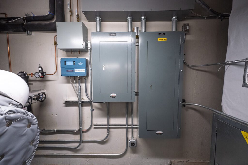 Three gray electrical panels and a blue control box are mounted on a beige wall, with metal conduit pipes running in multiple directions, indicating a utility or mechanical room setup that could benefit from Building Information Modeling for efficient management and maintenance.