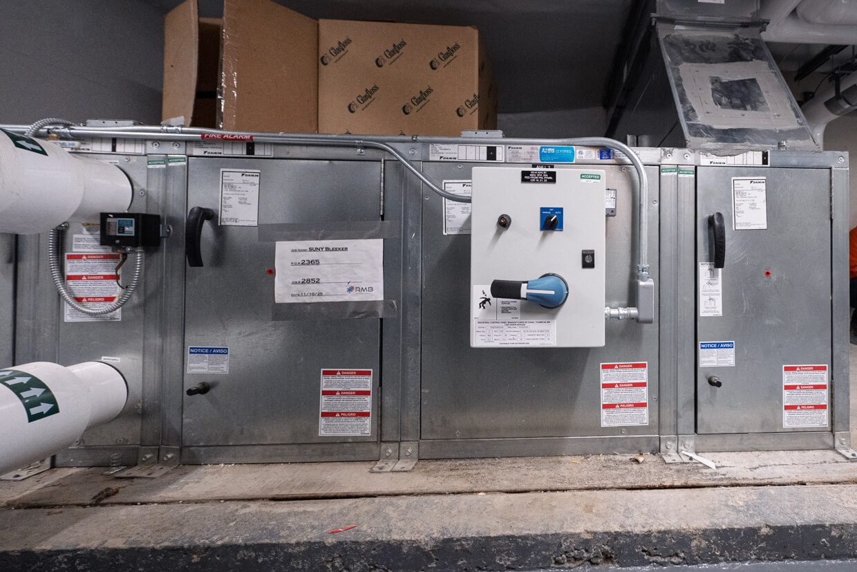 Large metal HVAC unit with control panel, pipes, and warning labels, situated in an industrial setting. Utilizing Building Information Modeling, the system is efficiently integrated within the space. Cardboard boxes are stacked on top in the background.