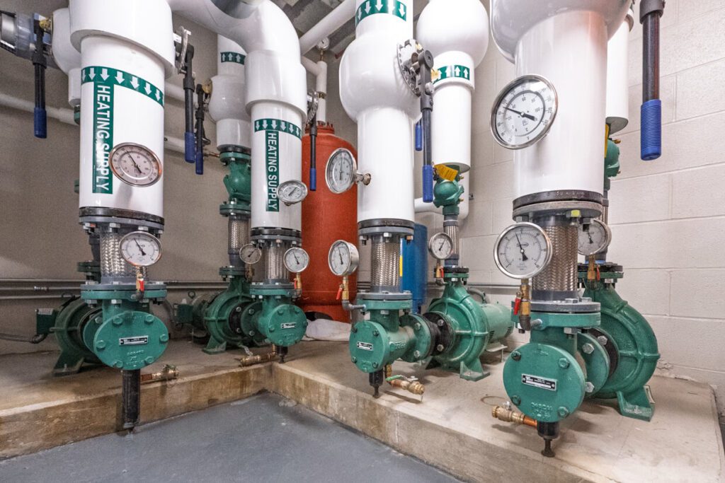 Four green heating supply pumps with pressure gauges and white insulated pipes are installed in a mechanical room. The organized layout, often designed using Building Information Modeling, features a red vertical tank in the background against a light-colored wall.