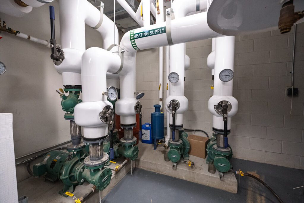 Several large white pipes labeled HEATING SUPPLY with gauges and green valves are connected to equipment in a utility room with a gray floor and white brick walls, illustrating how Building Information Modeling can streamline the design and management of complex mechanical systems.