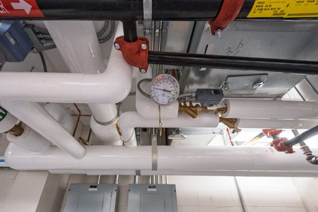 A network of white pipes with red fittings and a round pressure gauge, located on a ceiling in an industrial or utility room. Electrical panels and metal ducts are visible in the background, highlighting the organized infrastructure often detailed through Building Information Modeling.