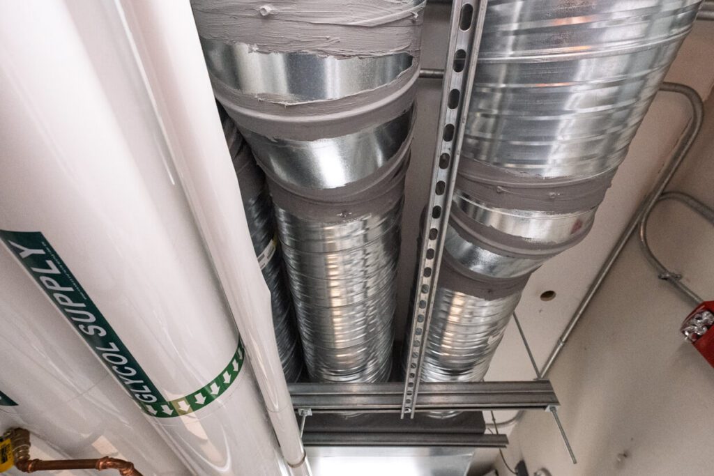 Large metal air ducts and a labeled Glycol Supply white pipe are suspended from the ceiling, with a metal support bar and a red fire extinguisher visible on the right side—an arrangement that exemplifies the organizational clarity made possible by Building Information Modeling.