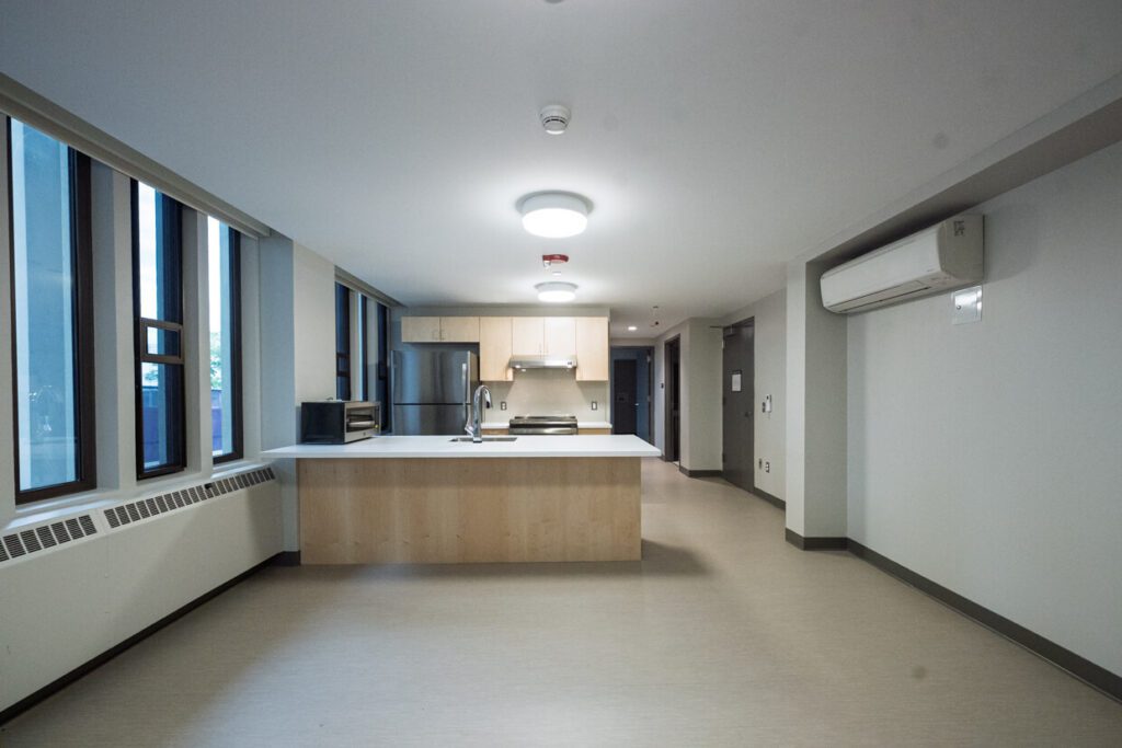 Modern, unfurnished apartment with large windows, kitchen island, stainless steel appliances, overhead lighting, white walls, and a wall-mounted air conditioning unit. Designed with precision using Building Information Modeling techniques, the space is open and clean with neutral flooring.