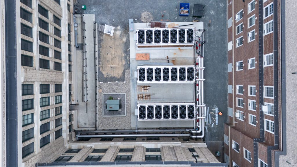 Aerial view of a building rooftop featuring large HVAC units, pipes, and gravel sections, surrounded by tall brick and concrete building walls—an ideal setting for integrating Building Information Modeling to streamline maintenance and management.