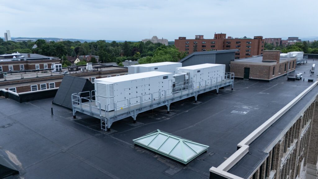 Large white HVAC units are mounted on a metal platform on a flat, black rooftop of a building, surrounded by brick structures and trees, with other buildings visible in the distance under a cloudy sky—a scene that exemplifies how Building Information Modeling can streamline the planning and coordination of complex rooftop installations.