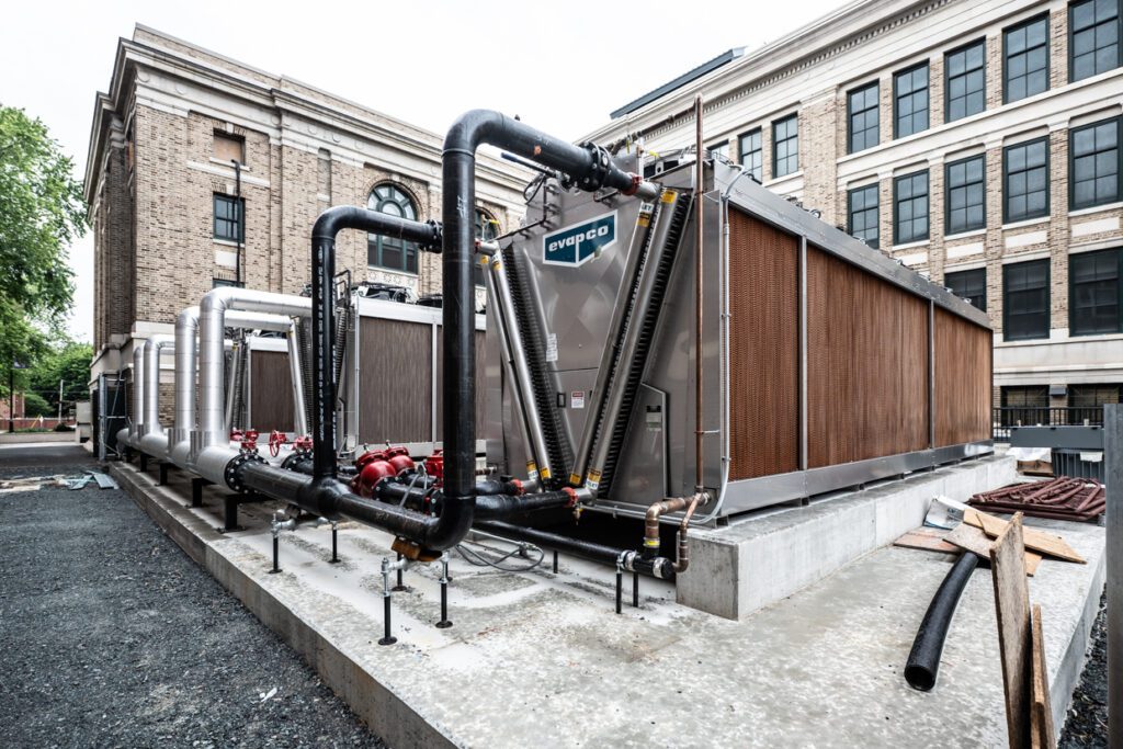 Large industrial HVAC cooling units with exposed pipes and pumps are installed on a concrete platform outside a multi-story brick building with large windows. Construction materials and gravel are visible nearby, highlighting a project that likely benefited from Building Information Modeling to coordinate such complex mechanical systems and site logistics.