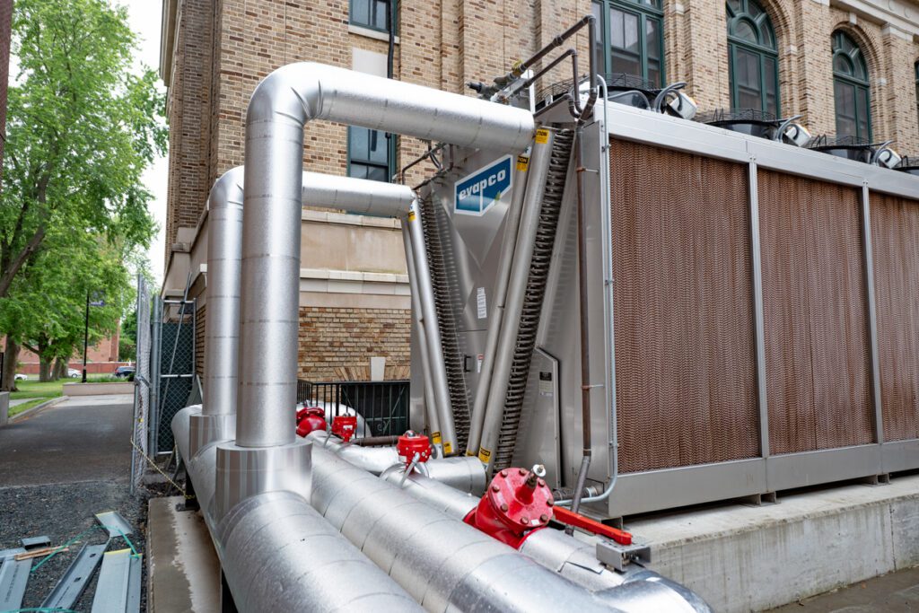 Large industrial HVAC unit with insulated metal pipes and red valves, installed outside a brick building, utilizing Building Information Modeling to ensure precise integration. Trees and a sidewalk are visible in the background.