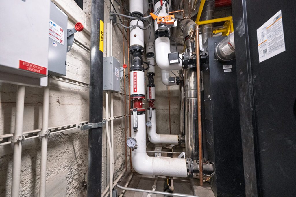 White insulated pipes with valves, pressure gauges, and labels are installed along a concrete wall in a mechanical room, surrounded by various control boxes and exposed wiring—a layout optimized through Building Information Modeling to enhance efficiency and organization.