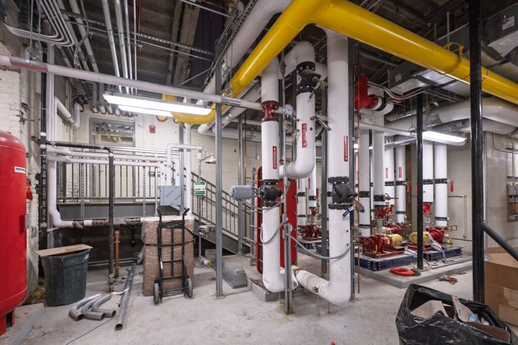 Industrial mechanical room with white and yellow pipes, red valves, gauges, and pumps. Stacks of cardboard boxes and a black trash bag are on the floor. Overhead, metal conduits and wiring are visible—a space that could greatly benefit from Building Information Modeling to streamline maintenance and facility management.