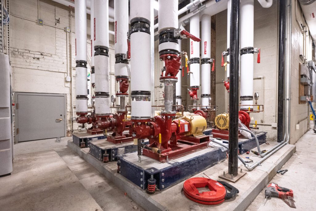 Large industrial pumps and red pipes with white insulation are installed in a mechanical room with concrete floors and walls. Tools and equipment lie scattered on the ground nearby, illustrating the ongoing installation process guided by Building Information Modeling for enhanced precision and efficiency.