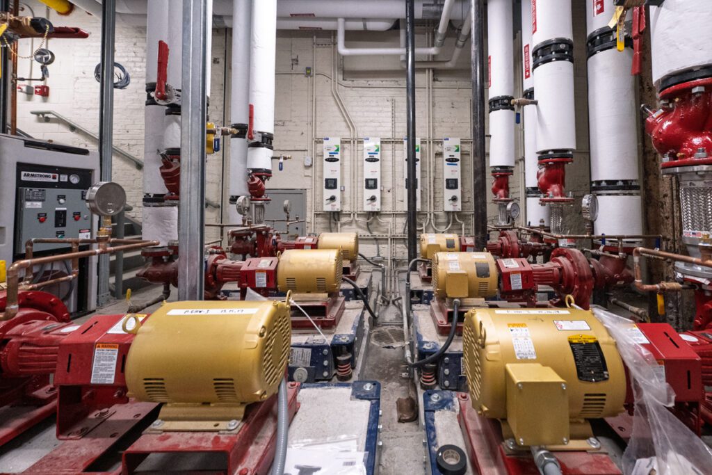A mechanical room with multiple red and yellow industrial pumps, pipes wrapped in insulation, electrical panels on the back wall, and various control boxes for building systems, all precisely organized using Building Information Modeling.