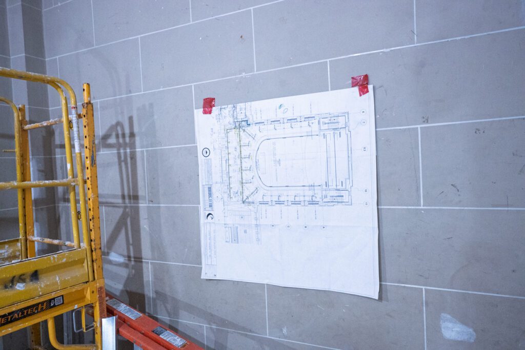 A construction blueprint, possibly created using Building Information Modeling, is taped to a tiled wall with red tape. Part of a yellow scaffold and an orange ladder are visible on the left side of the image.