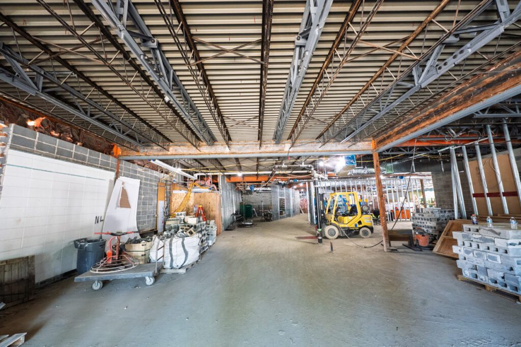Interior of a building under construction, with exposed steel beams, construction materials, a forklift, and unfinished walls and floors. Bright lighting illuminates the open, spacious area.