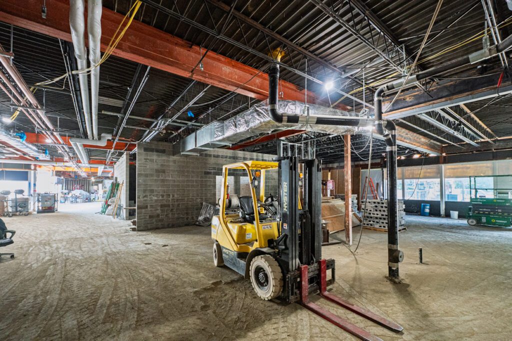 A yellow forklift is parked inside a large building under construction, with exposed beams, pipes, bricks, and construction materials visible throughout the area.
