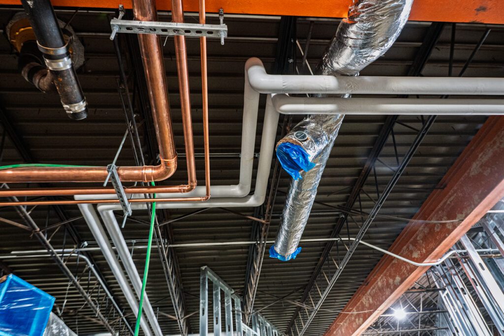 Pipes and air ducts run along an unfinished industrial ceiling, with insulation and blue tape on some ducts, and exposed metal beams visible.