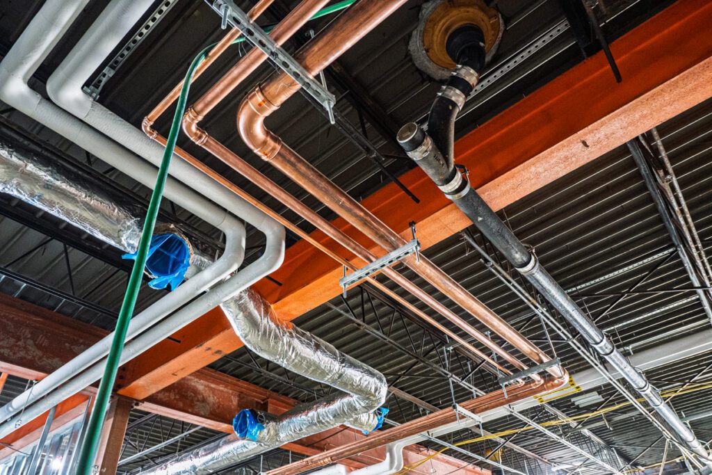 Exposed pipes, ducts, and wiring on an unfinished ceiling, including copper and black pipes, white PVC pipes, and silver air ducts wrapped in insulation, with some sections covered in blue plastic.