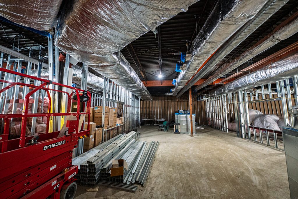 Interior of a building under construction with exposed metal framing, ductwork, insulation, a red lift, metal beams on the ground, and boxes stacked along the walls.