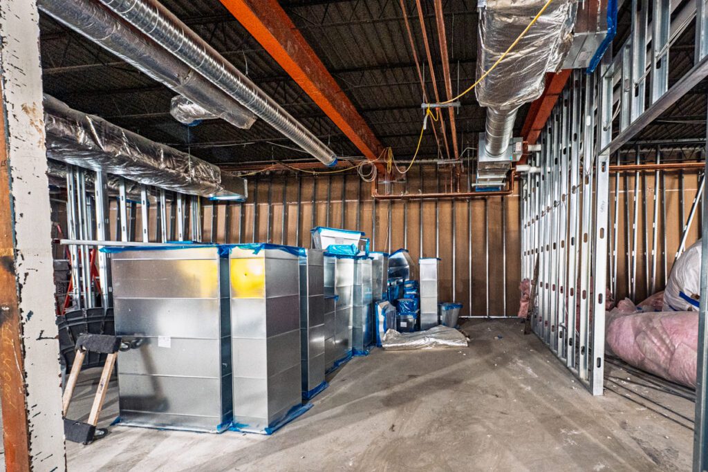 Large HVAC units in a partially constructed building with exposed metal framing, ducts, insulation, and wiring overhead. The floor is unfinished, and construction materials are scattered around the space.