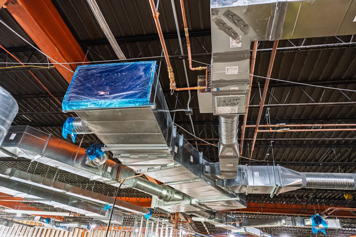 Exposed HVAC ductwork and metal pipes run along a building’s unfinished ceiling, with some sections covered in blue protective plastic and visible steel beams overhead.