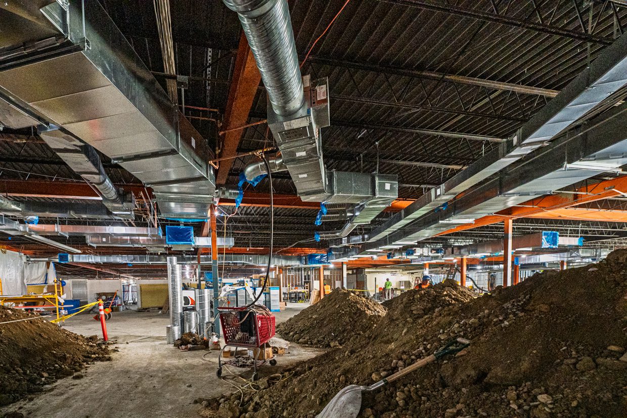 A large indoor construction site with exposed ducts, steel beams, and piles of dirt on the ground. Construction materials and tools are scattered throughout the area.