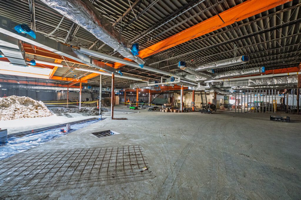 Large indoor construction site with exposed beams, ducts, and wires overhead. Piles of construction materials and equipment are scattered across the unfinished concrete floor.